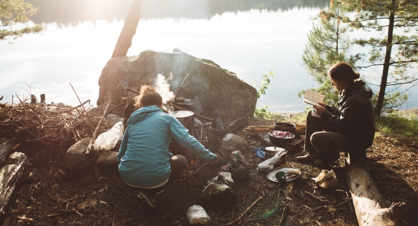 On the shore of a lake, one person tends to a campfire, while another sits nearby and reads. 